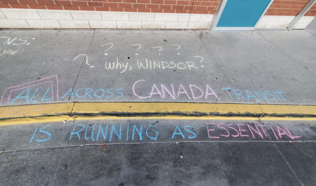 Colourful chalk writing on a sidewalk. It says "Why Windsor? All across Canada, transit is running as essential."