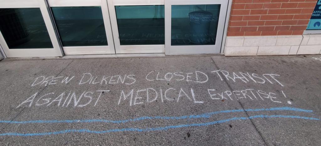 "Drew Dilkens closed transit against medical expertise!" written in chalk across a sidewalk in front of a building.