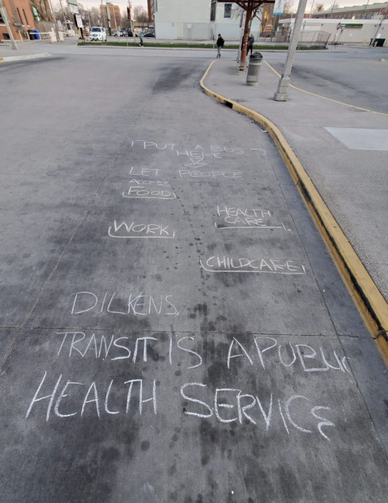 Chalk writing at a bus stop. It says "Put a bus here: Let people access food, healthcare, work, childcare. Dilkens, transit is a public health service."