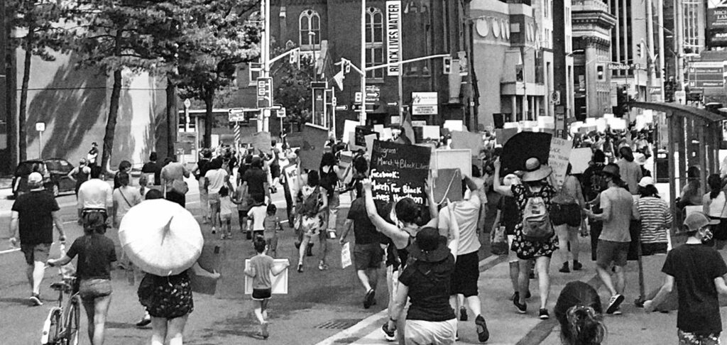 Black and white photo of marchers in Hamilton, with their backs turned to the camera, holding Black Lives Matter signs. A nearby building has a large "Black Lives Matter" banner hanging from its walls.