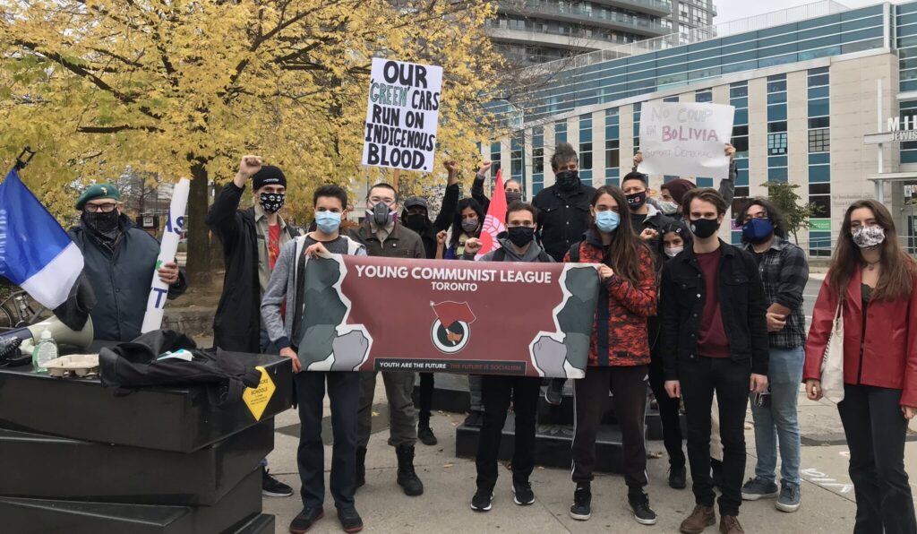 A group of young protesters gathers in Toronto. Most are wearing autumn jackets, and they are on a street corner. A group holds up the banner of the Young Communist League, and some hold up anti-coup signs. One older protestor holds the MAS Party flag.