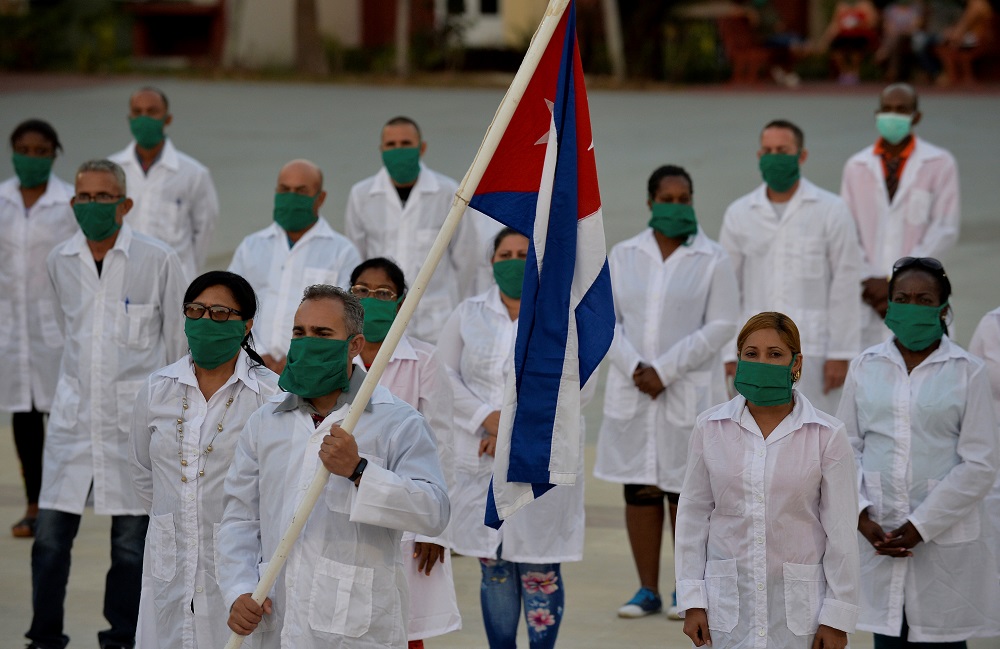 Cuban medical workers in white coats and green face masks hold the Cuban flag. They are standing in a group of around 15 people and look towards the sky.