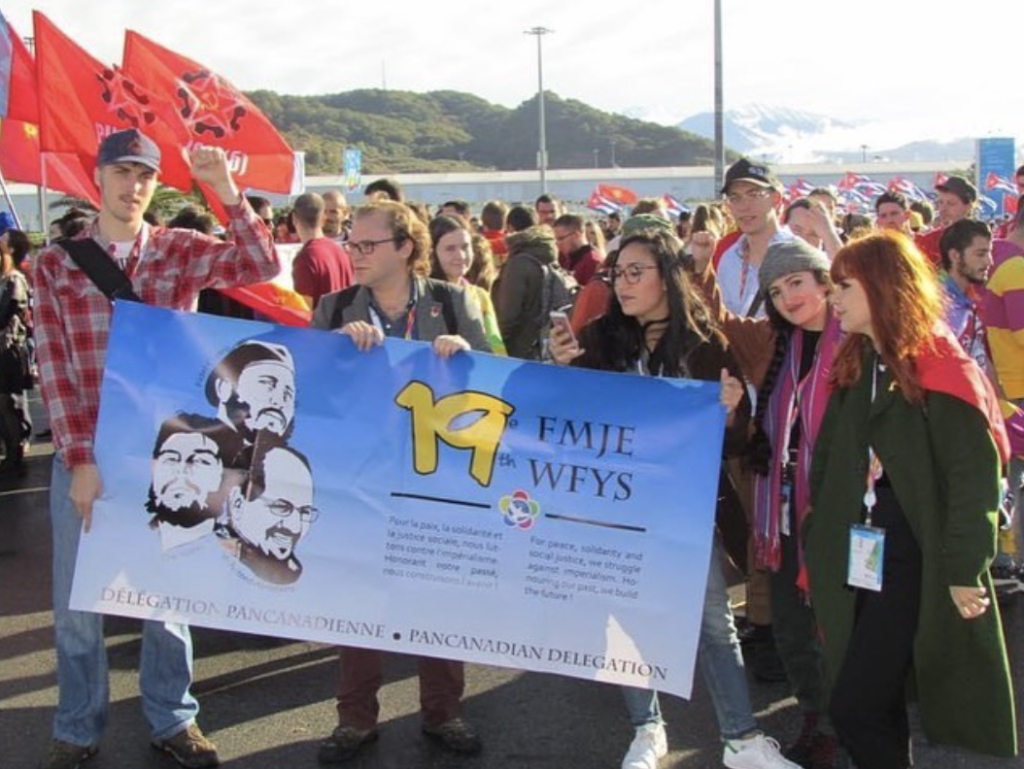 Five YCLers hold up the banner of the Pan-Canadian Delegation at 2017's World Festival of Youth and Students in Sochi. General Secretary Ivan Byard raises his fist. The banner is blue and reads "19th WFYS/19e FMJE: Délegation Pancanadienne" Behind them are many young people, most holding red flags. Also in the background are a large group of Cuban and Vietnamese flags and a mountain range.