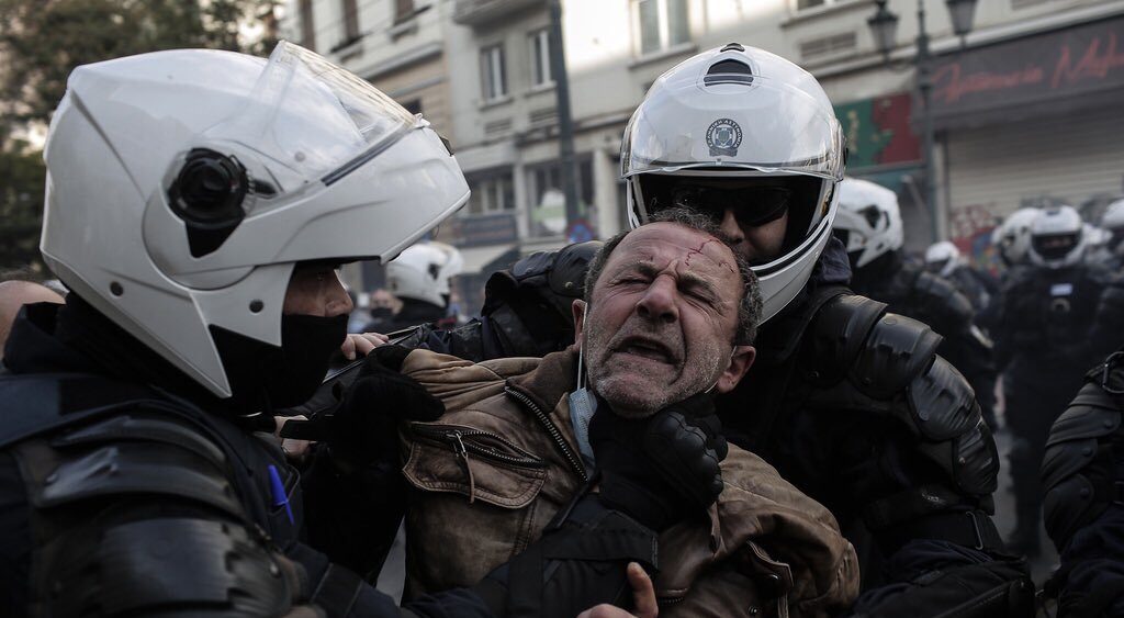 Greek police grab an older man by the neck, pulling down his mask.