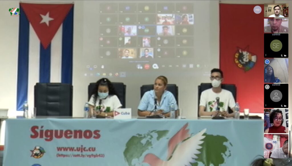 A screenshot of a video call shows three Cuban representatives sitting at a desk. Behind them are the Cuban flag and a UJC flag, as well as a projection of the small thumbnails of other participants. The table they sit at has a picture of the conference logo, a dove and a globe.