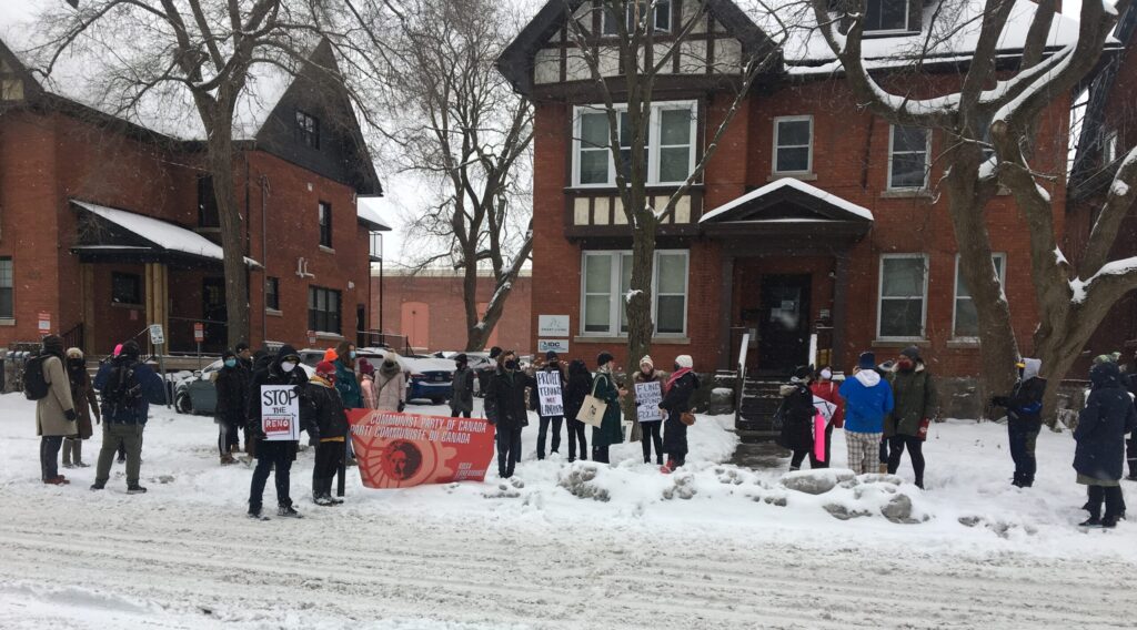 A group of people stands in a snowy street in front of some red brick townhouses. Some members of the group are holding a Communist Party banner, and others have anti-renoviction signs.