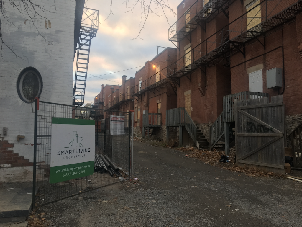 A photo of a fenced-off alley with a row of red brick apartments, with all windows and doors boarded up. There is a sign that says "Smart Living Properties".