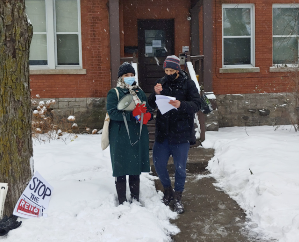 Two people wearing masks and winter coats stand in the snow. There is a sign on the ground that says "Stop the Reno". One of the people, Doug, is holding a few sheets of paper that he is reading from. The other person, Michelle, holds a megaphone.