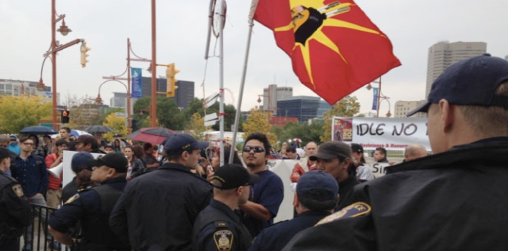 A man waves the Mohawk Warrior flag while being confronted by police at the opening of the Canadian Museum for Human Rights. An Idle No More banner is visible in the background.