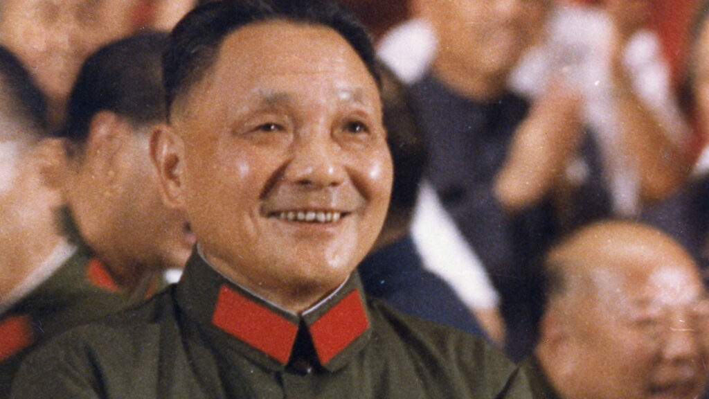 Deng Xiaoping smiles. He is wearing a green shirt with red lapels. Behind him you can see a crowd of clapping people.