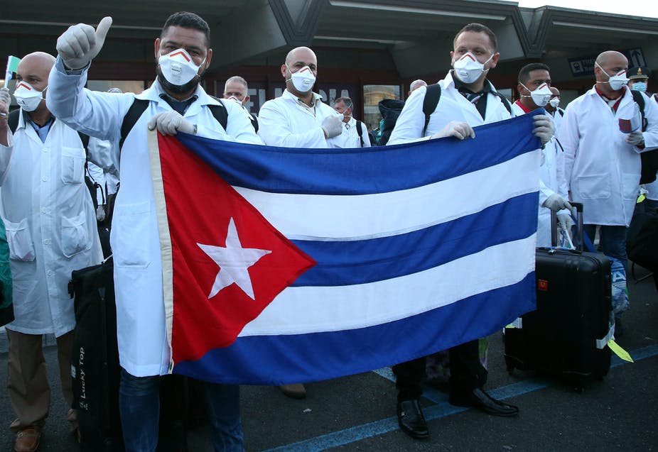 A group of Cuban doctors in white coats and masks hold the Cuban flag. One is looking at the camera and giving a "thumbs up" gesture.