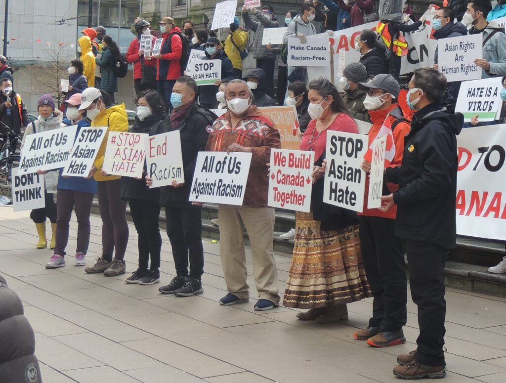 A large group of Indigenous and East Asian people hold up signs that say "Stop asian hate", "We build Canada together", and "end racism" at a rally in Vancouver.