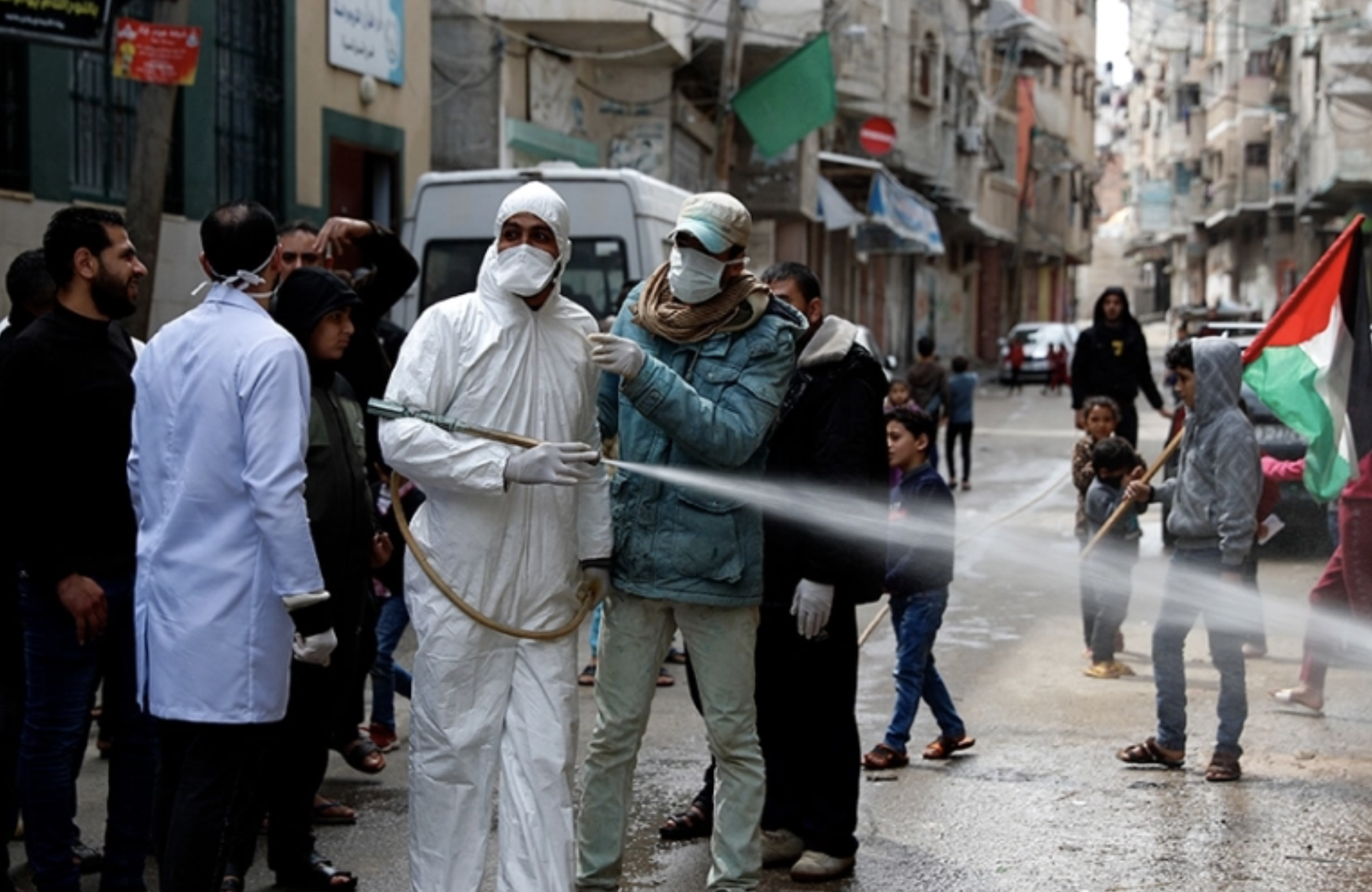 Men in hazardous material suits and surgical masks spray sanitizer in the crowded Gaza strip. a Boy with a Palestinian flag can be seen walking in the background.