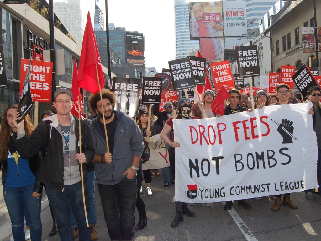 Young communists carry a banner outside of Ryerson University in Toronto that says "Drop fees, not bombs". Others carry red flags or signs that say "Free education now" and "fight the fees". They make up a very large group of people and are marching in the middle of the street towards the camera.