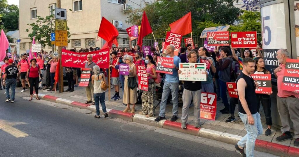 A large group of Israeli protesters gathers on the street. They hold signs in Hebrew and Arabic that say slogans like "Jews and Arabs refuse to be enemies". The signs are red, and some protesters hold red flags.