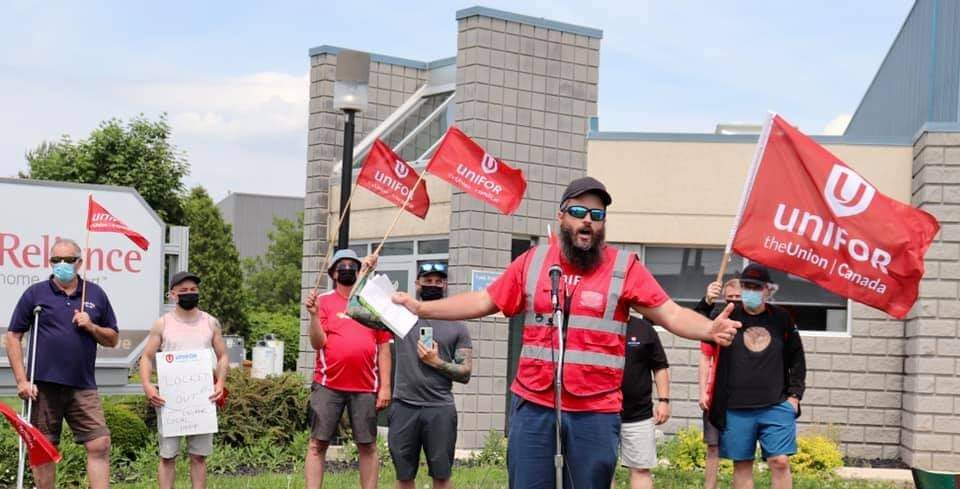 A man in a red safety vest outstretches his arms and speaks into a microphone. Behind him is a crowd holding red Unifor union flags. There is a building with a sign that says "Reliance" behind the crowd.