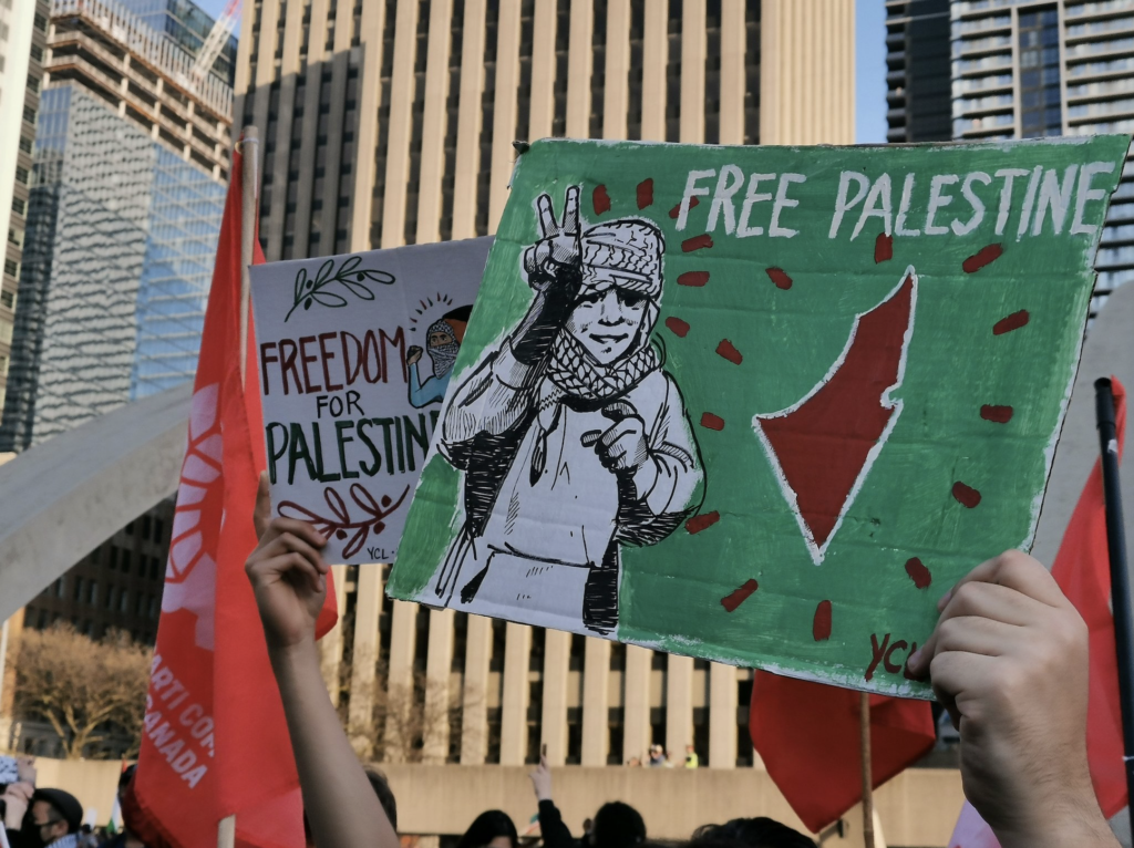 Two YCLers hold signs at a rally. One says "Free Palestine" on a green background, with a drawing of a child wearing a keffiyeh and flashing a peace sign. The green sign has an outline of Palestine. The other sign is on a white background and says "freedom for palestine" with olive leaves. Behind the signs is a Communist Party flag and some urban buildings.
