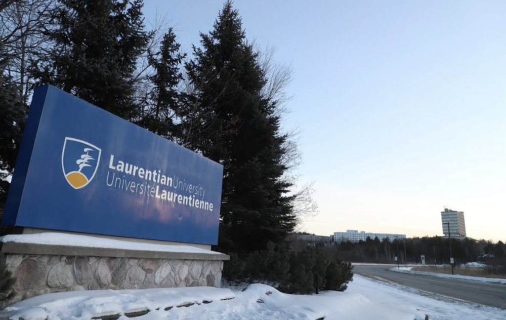 A large blue Laurentian University sign stands amongst snow and pine trees.
