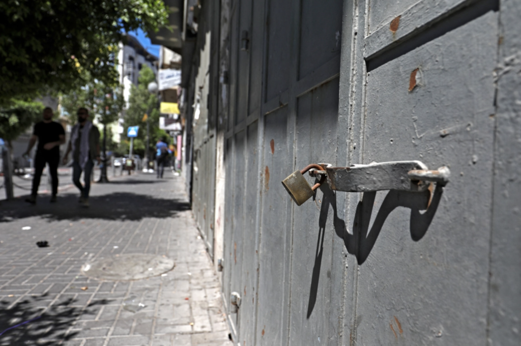 Photo depicts a lock on a grey door. In the background you can see some people walking. The photo was taken in Israel or occupied Palestine and there are green trees and a dusty street in the background.