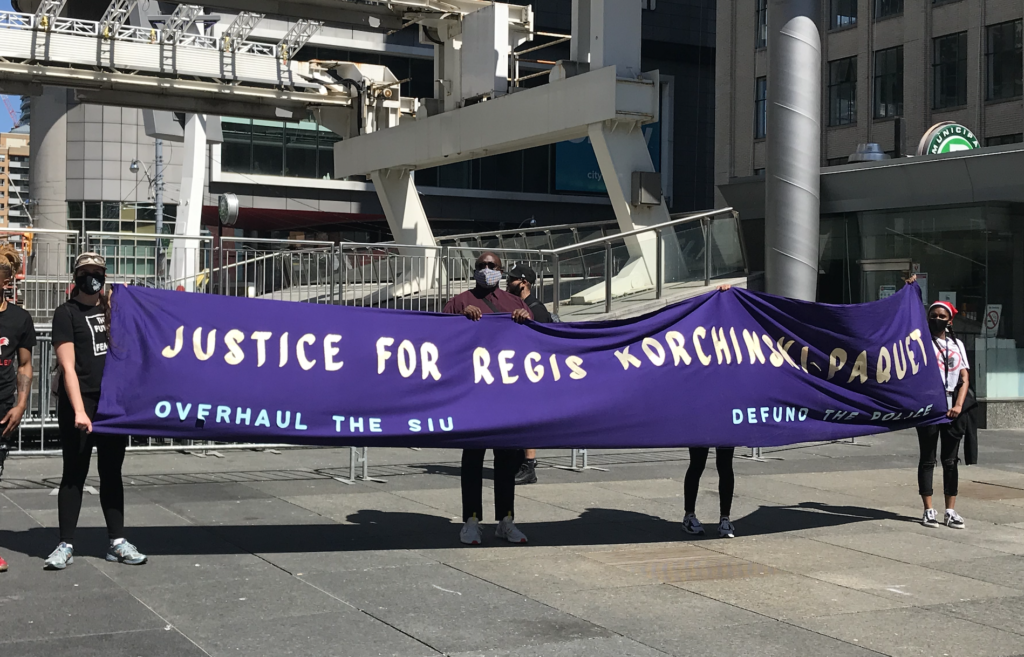 A group of marchers holds a long purple fabric banner that says "Justice for Regis Korchinski-Paquet"