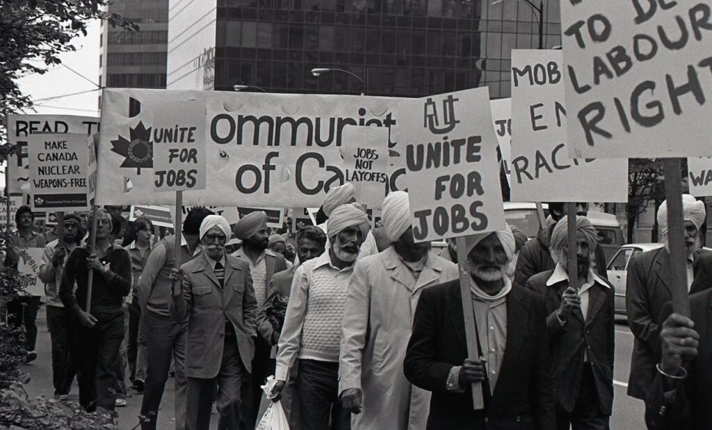 A black and white photo from the 1980s shows a large group of Sikh Communist Party of Canada members marching and holding banners. The banners show slogans like "unite for jobs" or simply the name of the Communist Party.