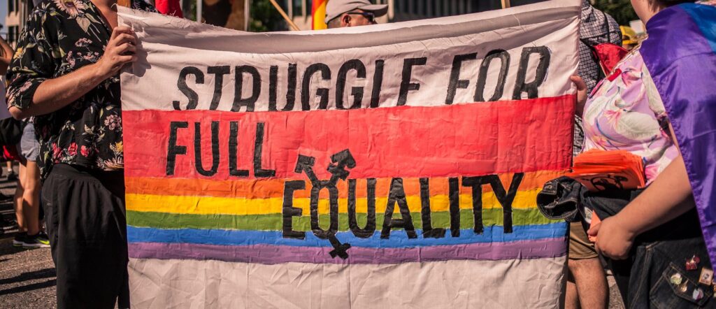 A painted banner with a pride rainbow. The red of the rainbow is larger than all other stripes. On top of the rainbow it says "Struggle for full equality". The Q of equality is a transgender symbol.