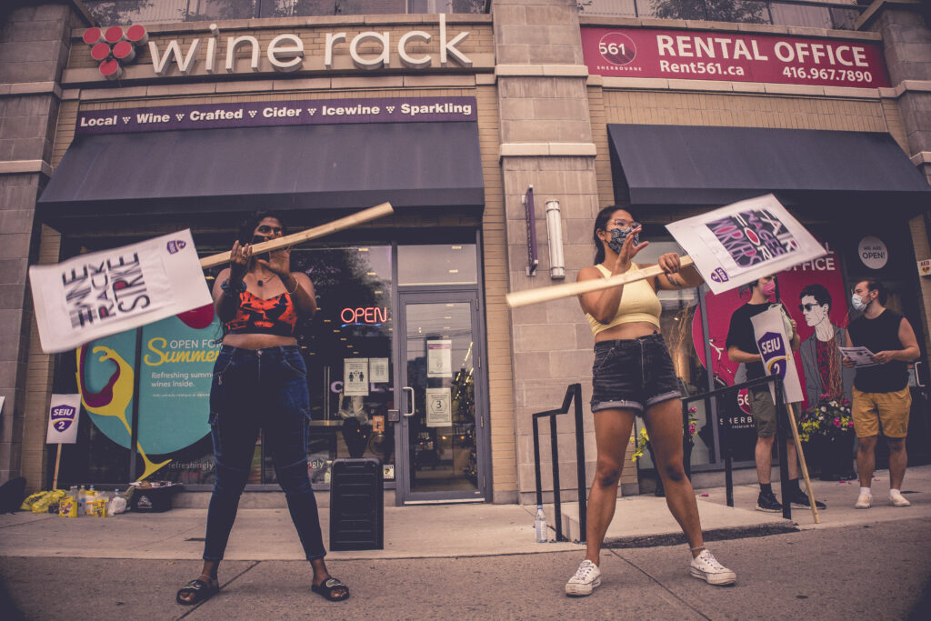 Two Wine Rack workers twirl protest signs outside of a Wine Rack store. In the background, a YCLer and a Wine Rack worker discuss the strike. The signs say "SEIU 2", "FAIR WAGES FOR WORKERS", and "WINE RACK STRIKE"