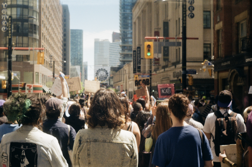 A large group of young people gathers at Toronto's Yonge and College intersection. The sky is blue and the group is surrounded by tall buildings and street lights. Some hold signs that say "ACAB", and they all face away from the camera.