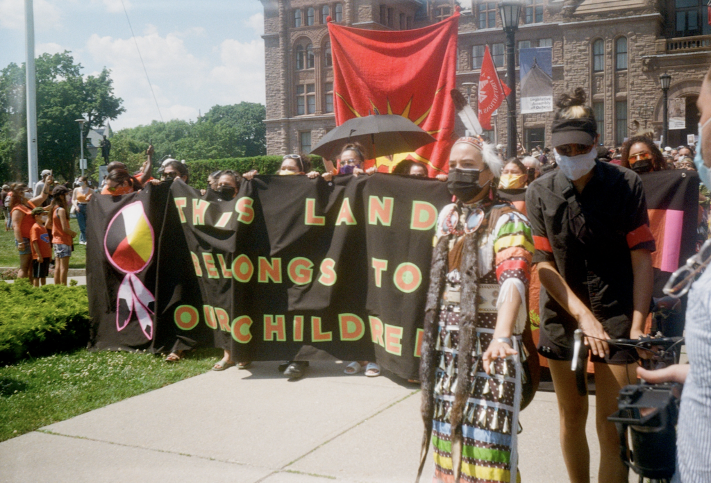 A group of Indigenous people in cultural dress leads a procession out of Queen's Park, the Ontario seat of parliament. They are holding a banner that says "This land belongs to our children". One woman in the front gestures with her hands to have attendees move back.