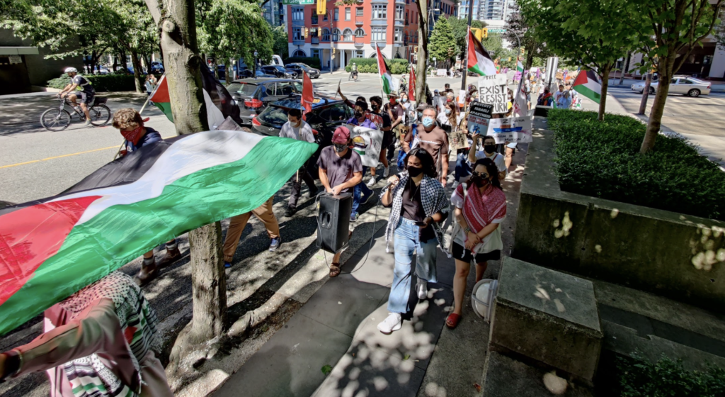 A large group of people wearing keffiyeh (Palestinian traditional scarves) walks down a street. A large Palestinian flag is carried horizontally by multiple people.