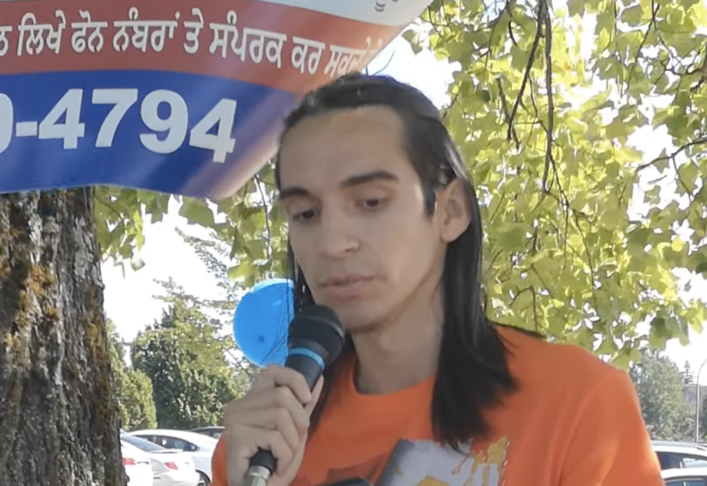 Alex speaks into a microphone, wearing an orange shirt. A park with trees is shown behind them.