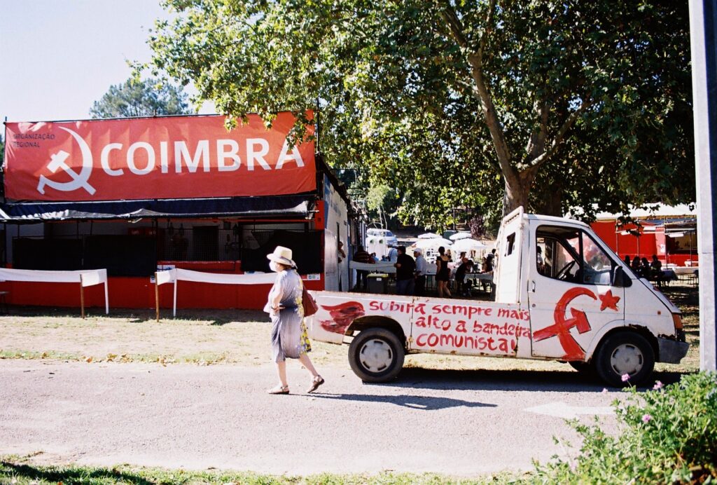 A photo shows a woman walking down a path at the Festa do Avante! in Seixal, Portugal. Behind her is a booth for the Coimbra region with a large hammer and sickle. Beside her is a truck with a hand-painted message and hammer and sickle, reading "We will always raise the communist flag higher" in Portuguese.