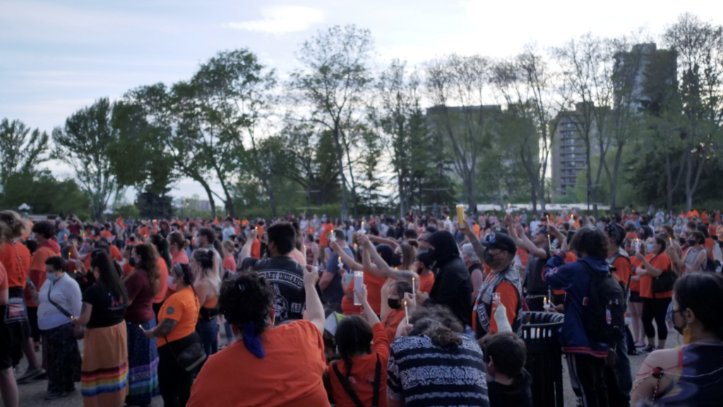 A large group of people wearing orange shirts gathers in Edmonton. It is dusk and the photograph depicts hundreds of people facing away from the camera.
