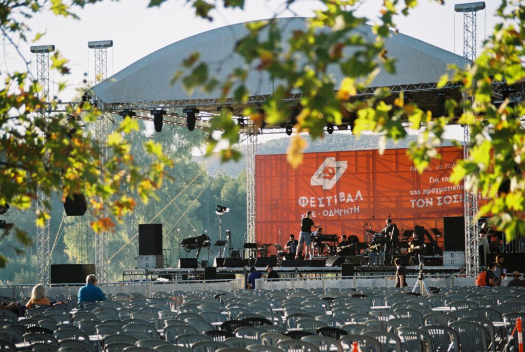 A photo of a sunny concert stage, surrounded by trees and mountains. The stage backdrop is red, with a hammer and sickle.