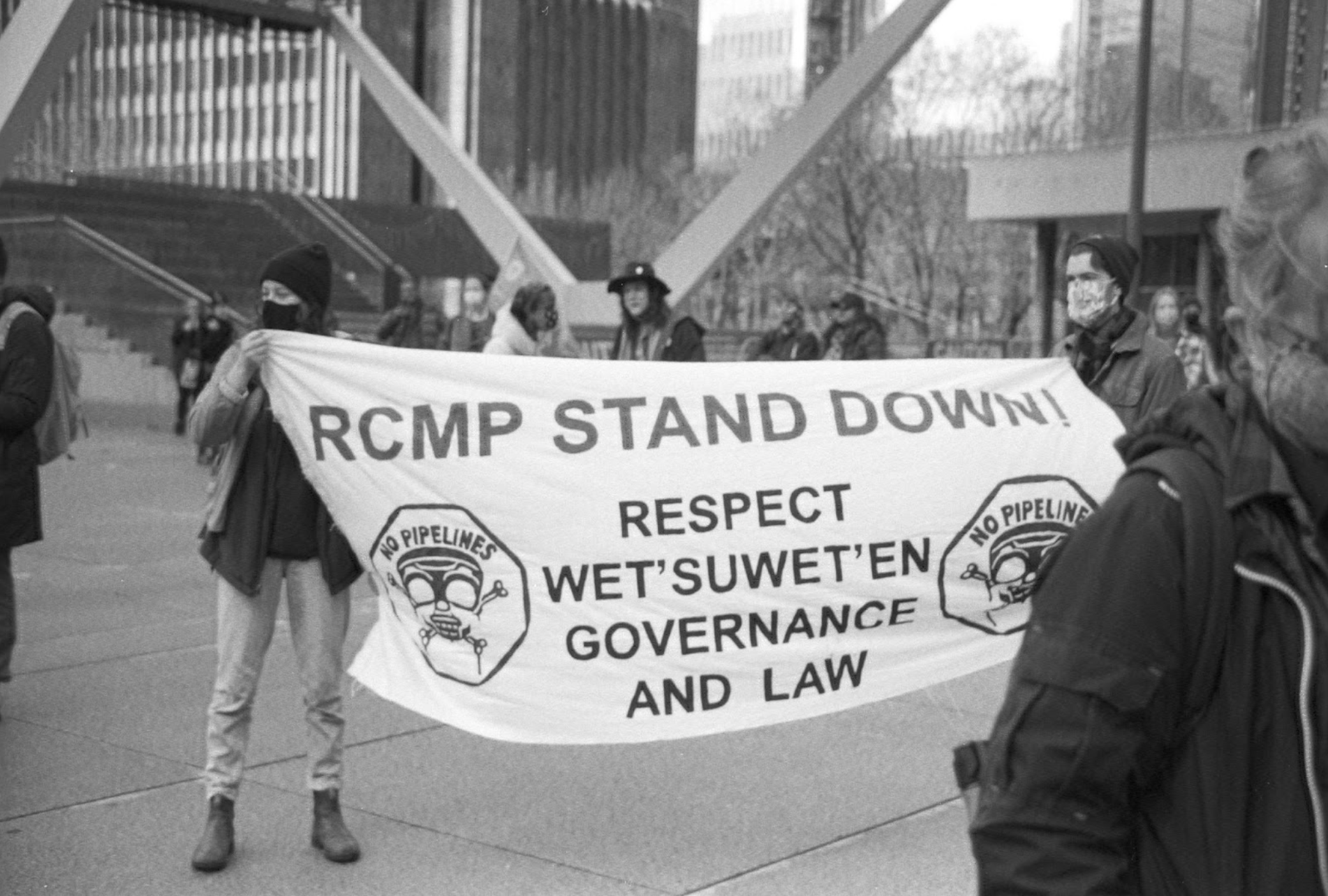 A black and white image of two people holding a large white banner. The banner reads "RCMP stand down! Respect Wet'suwet'en governance and law" in large capital letters. Behind them you can see an urban landscape.